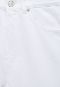 Jean en denim blanc présentant une texture lisse, un design à cinq poches et un bouton métallique avec insigne de la marque.