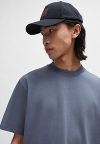 Tricou gri cu mânecă scurtă, cu logo brodat; textură moale. Casca neagră cu patch logo pe față; vizieră structurată, stil casual.