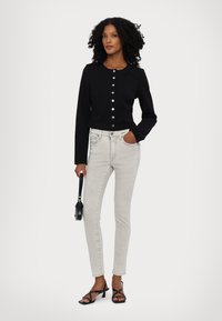 Czarna dopasowana bluzka z długim rękawem i srebrnymi guzikami, w połączeniu z jasnoszarymi rurkami i czarnymi sandałkami na szpilce, trzymająca czarną torebkę.