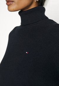 Donna che indossa un maglione a collo alto a coste nero con una piccola toppa con logo rosso, bianco e blu sul petto, e orecchini dorati.