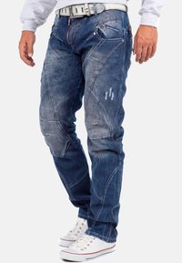 Jeans in denim blu con una vestibilità slim, cuciture a contrasto e dettagli strappati. Indossati con sneakers bianche e una felpa grigia.