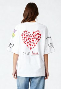 Balta per didelė t-shirt su raudonų širdelių raštu nugaroje, užrašas "SWEET heart" ir gėlių grafika ant rankovių. Šviesūs džinsai.