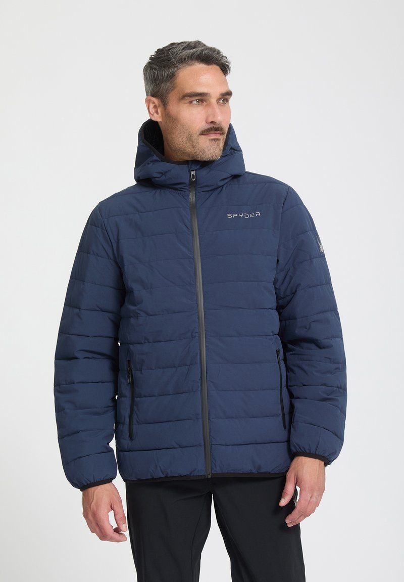 Homme portant une veste matelassée à capuche bleu marine avec fermeture éclair devant et poches latérales, regardant vers sa gauche sur un fond uni.