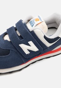 Scarpa sportiva in suede e mesh blu navy con dettagli bianchi, cinghie in velcro, suola rossa e logo visibile sul lato. Progettata per il comfort.