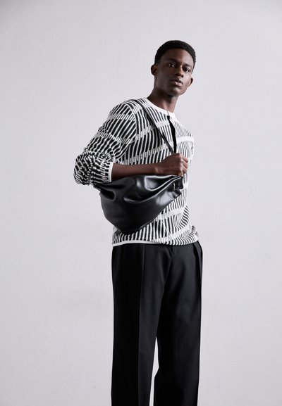 Μαύρη δερμάτινη τσάντα με γυαλιστερή επένδυση, ριγέ μαύρο-άσπρο πλεκτό πουλόβερ και χαλαρές μαύρες παντελόνες. Το μοντέλο ποζάρει μπροστά σε απλό φόντο.