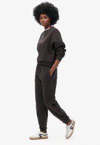 Sudadera y pantalones deportivos marrón oscuro hechos de tela suave, con cintura ajustable con cordón. Zapatillas con detalles en blanco y negro, suela texturizada.