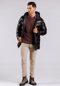 Chaqueta acolchada marrón con capucha azul, usada sobre un suéter burdeos texturizado y pantalones beige. Botas marrones completan el atuendo.
