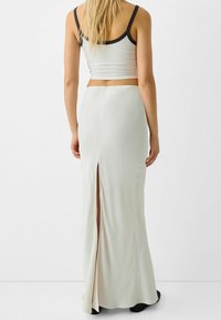 Žena v bílé maxi sukni s vysokým pasem a rozparkem na zadní straně, doplněná bílým krátkým tílkem s černými lemy.