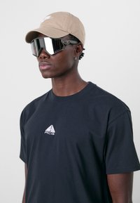 Schwarzes T-Shirt mit aufgesticktem ACG-Logo, beiger Kapuzenmütze und reflexionsstarken übergroßen Sonnenbrillen mit schwarzen Akzenten. Glatte Texturen und modernes Design.