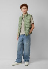 Jasnozielona pikowana kamizelka na beżowym t-shircie, w połączeniu z luźnymi niebieskimi dżinsami i białymi trampkami. Gładka tekstura, casualowy design.