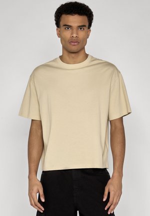 Jeune homme aux cheveux courts et bouclés, portant un t-shirt beige ample à manches courtes et un pantalon noir, debout devant un fond uni.