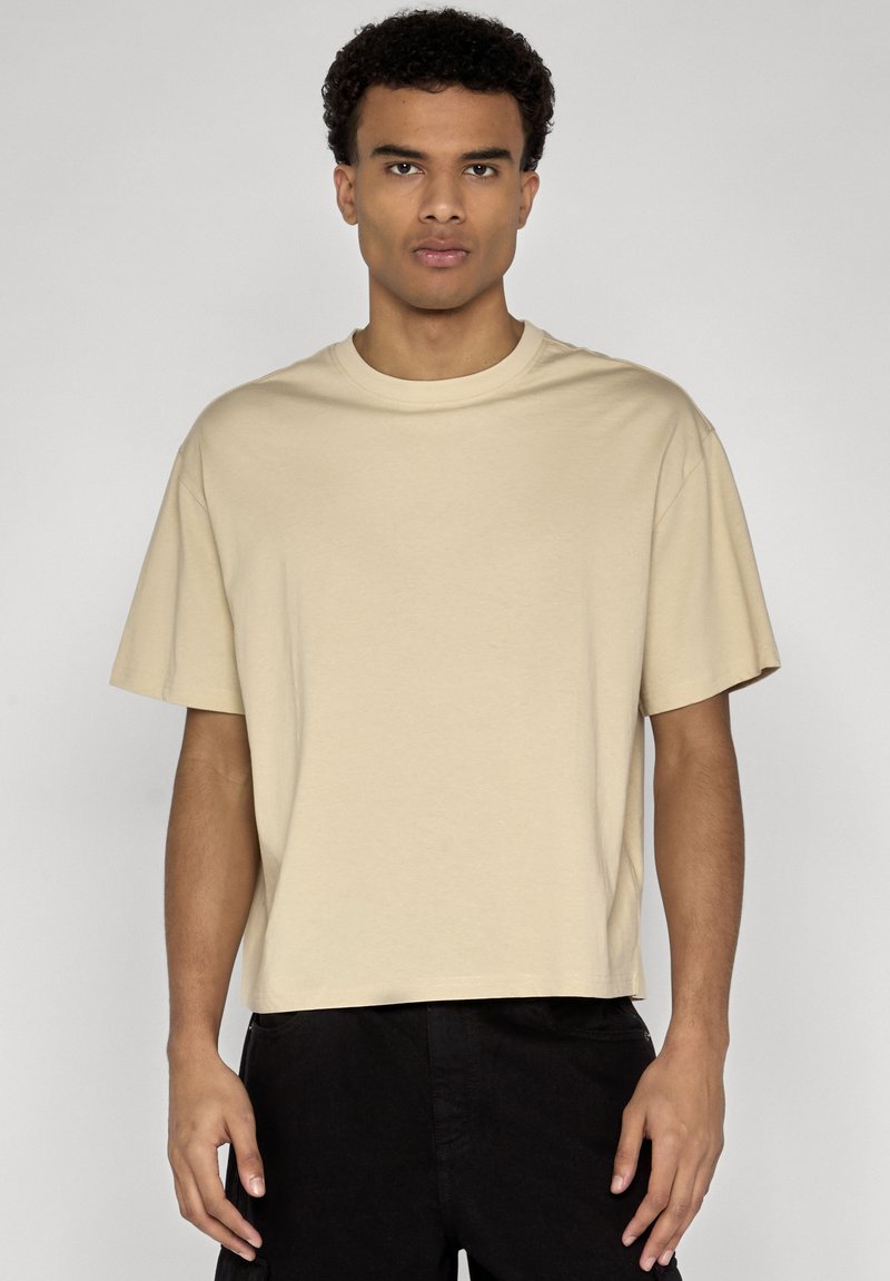 Jeune homme aux cheveux courts et bouclés, portant un t-shirt beige ample à manches courtes et un pantalon noir, debout devant un fond uni.