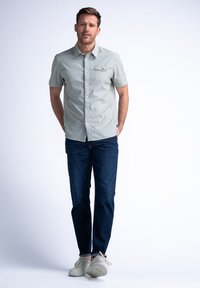 Camisa de botones de manga corta, verde claro, con un estampado sutil, combinada con jeans azul oscuro y zapatillas de colores claros.