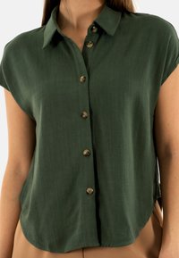 Camicia a maniche corte, verde oliva, con bottoni in tessuto testurizzato. Ha tre bottoni e colletto ripiegato. Vestibilità casual e ampia.