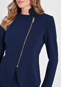 Veste ajustée bleu marine avec un col haut, dotée d'une fermeture éclair diagonale en or et de coutures structurées. Tissu lisse avec une silhouette moderne.