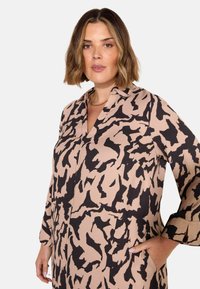 Blusa a maniche lunghe in beige chiaro con un motivo astratto nero, caratterizzata da un colletto e tasche laterali. Tessuto morbido con una vestibilità comoda.