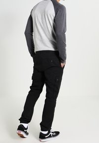 Man med händerna i fickorna iförd en grå och mörkgrå långärmad tröja, svarta cargo-byxor, vita strumpor och svarta sneakers.