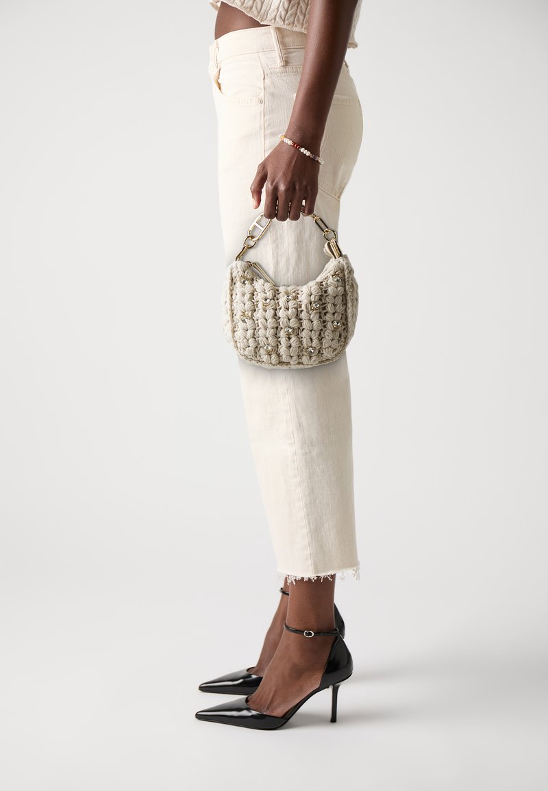 Knit handbag in light beige with textured surface and metal accents, carried by a person in cream jeans and black pointed-toe heels.