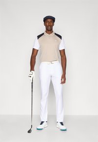 Pólo bege e branco com detalhes em azul marinho, calças brancas, luvas de golfe e sapatos. Pose em pé segurando um taco de golfe.