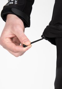 Vêtement noir avec un poignet et un cordon élastique ajustable. Une main tient le cordon, mettant en avant une texture lisse et un design épuré.