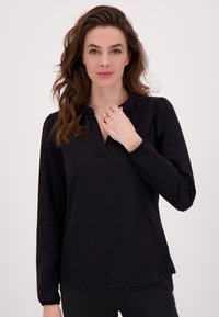 Vrouw met lang golvend bruin haar, gekleed in een zwarte blouse met lange mouwen, houdt haar hand bij haar hals en poseert tegen een effen witte achtergrond.