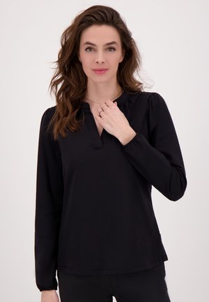 Vrouw met lang golvend bruin haar, gekleed in een zwarte blouse met lange mouwen, houdt haar hand bij haar hals en poseert tegen een effen witte achtergrond.