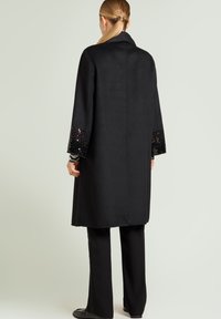 Svart yllekappa med rak skärning och långa ärmar; muddar prydda med svarta paljetter och metalliska accenter för textur. Matchas med svarta byxor.