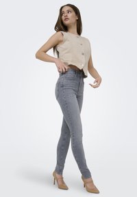 Femme portant un haut sans manches beige boutonné, un jean skinny gris et des chaussures à talons hauts beige, posant et regardant vers le haut sur le côté.