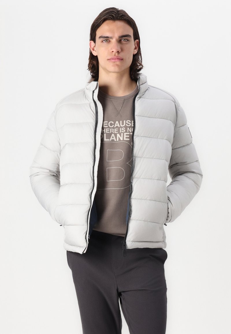 Chaqueta de abrigo ligera en color gris claro, con acolchado horizontal, cuello alto y cremallera. Llevada sobre un suéter marrón con diseño de texto. Bajo ajustable.