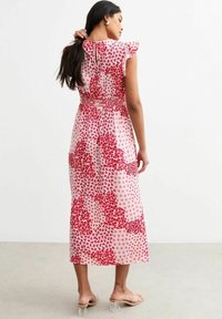 Vestido maxi con una base rosa y patrones florales rojos, mangas con volantes y detalle de cintura fruncida. Sandalias de tacón transparente complementan el look.
