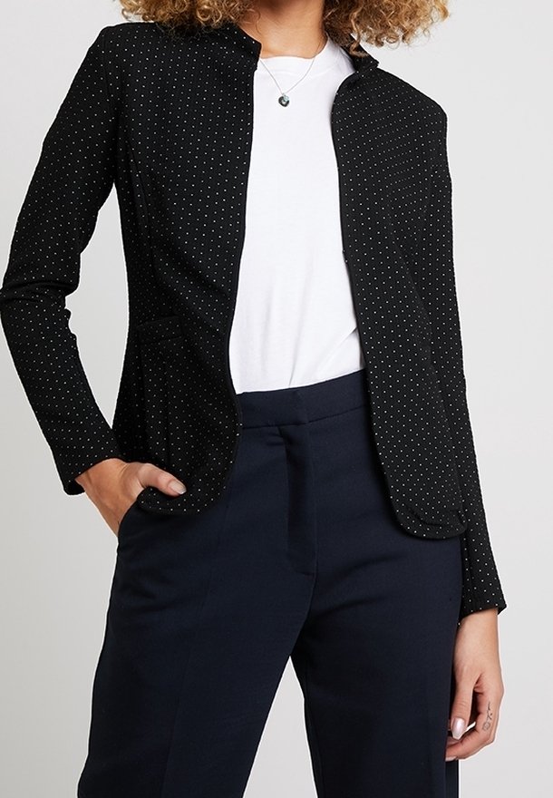 Femme portant un blazer noir à petits pois blancs, une chemise blanche, un pantalon bleu marine foncé et un collier argenté avec un pendentif.