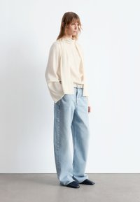 Leichtes cremefarbenes Oberteil mit hohem Kragen, lockerer Passform und voluminösen Ärmeln, kombiniert mit lockeren hellblauen Jeans und spitzen marineblauen Schuhen.