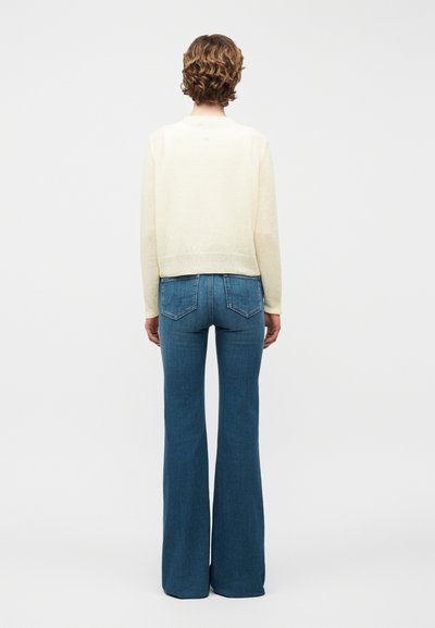 Άτομο με κοντά σγουρά μαλλιά που φοράει ένα κρεμ πουλόβερ και ψηλόμεσα μπλε τζιν με φαρδιά μπατζάκια, φαίνεται από πίσω σε απλό φόντο.