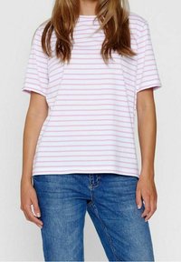 Žena v bílém tričku s tenkými růžovými vodorovnými pruhy a modrých džínách, stojící na jednobarevném bílém pozadí.