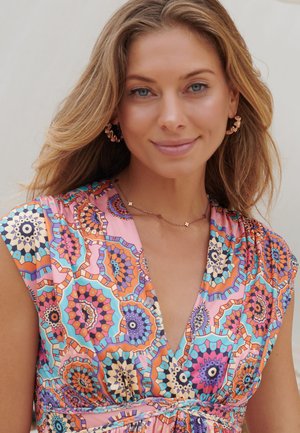 Lächelnde Frau mit langen blonden Haaren, die goldene Ohrringe und ein buntes ärmelloses Kleid mit geometrischen Mandala-Mustern trägt.