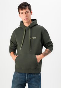 Sudadera verde oscura de tela suave, con un bolsillo frontal, cordones y logotipo bordado. Llevada con jeans azules.