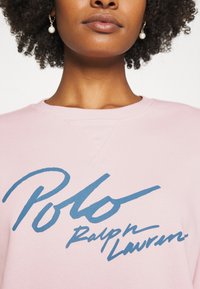 Ljust rosa sweatshirt med en stor blå "Polo Ralph Lauren"-logotyp. Tyget verkar ha en textur med rundad halsringning och synlig sömnad.