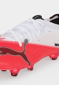 Λευκό και κόκκινο ποδοσφαιρικό παπούτσι Puma με λευκά κορδόνια και μαύρο λογότυπο Puma στο πλάι, σχεδιασμένο για χρήση σε σκληρό έδαφος.