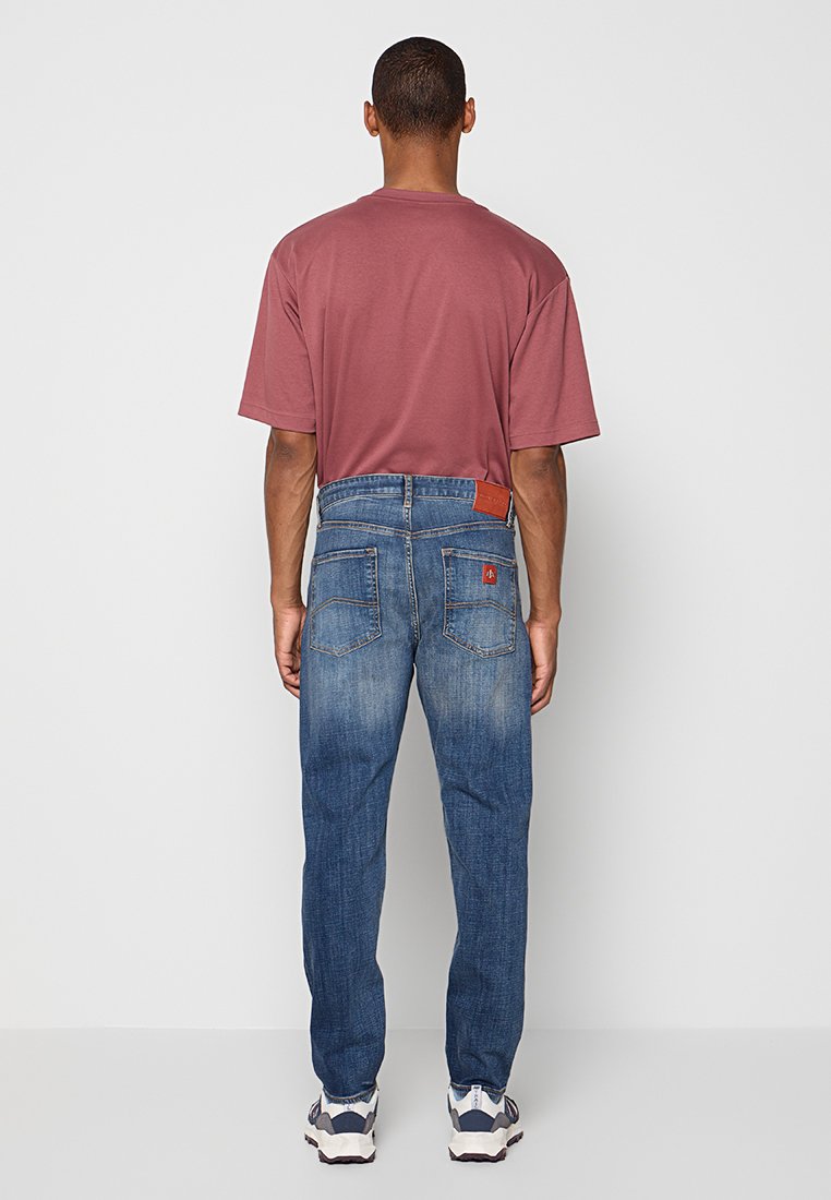 Uomo che indossa jeans blu sbiaditi, una maglietta oversize color marsala e sneakers bianche, in piedi di fronte a un muro grigio chiaro uniforme.