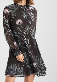 Schwarzes Kleid mit Blumenmuster in Pastellrosa und Blau. Hergestellt aus leichtem Stoff, mit langen Ärmeln und einem gerüschten Ausschnitt.