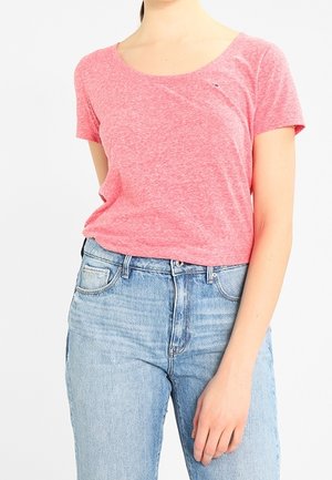 Kobieta ubrana w różowy melanżowy T-shirt z krótkim rękawem, włożony w wysokowalne jasnoniebieskie dżinsy, stojąca z jedną ręką opartą w pobliżu biodra.