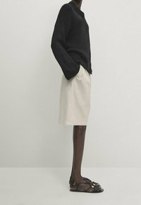 Musta neulottu neuletakki, kerma värinen polvipituinen hame, ruskeat litteät sandaalit, joissa on ременные yksityiskohdat, sivusta kuva, joka esittää rentoa istuvuutta ja erottuvia tekstuureja.