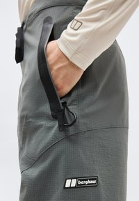 Main dans la poche d'un pantalon de plein air gris Berghaus avec une fermeture éclair noire et un clip, porté avec un haut beige à manches longues.