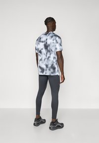 Herren-Hemd mit kurzen Ärmeln im schwarz-weißen Batikmuster, kombiniert mit enganliegenden, dunkelgrauen Leggings und schwarzen Sportschuhen.