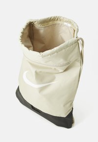 Sac à cordon beige clair avec un fond noir. Il présente un logo blanc, une texture lisse et un intérieur spacieux avec une ouverture renforcée.