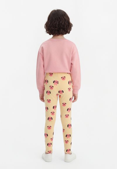 Dítě stojící zády, oblečené v růžovém tričku s dlouhým rukávem a žlutých legínách s růžovými a černými psími tvářemi a bílých botách.