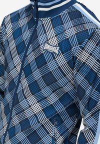 Mėlynos langelinės striukės, pagamintos iš lengvo audinio, su užtrauktuku priekyje, dryžuotu apykakle ir logotipo pleistru. Dizainas apima įvairias mėlynas atspalvius.