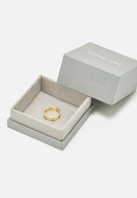 Anello d'oro con design circolare e quattro piccoli pietre trasparenti, adagiato su un morbido interno beige di una scatola argentata etichettata "Michael Kors".