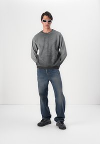 Grå sweatshirt med præget ørnedesign, parret med blå jeans. Modellen bærer hvide solbriller og sorte sneakers. Afslappet sæt.