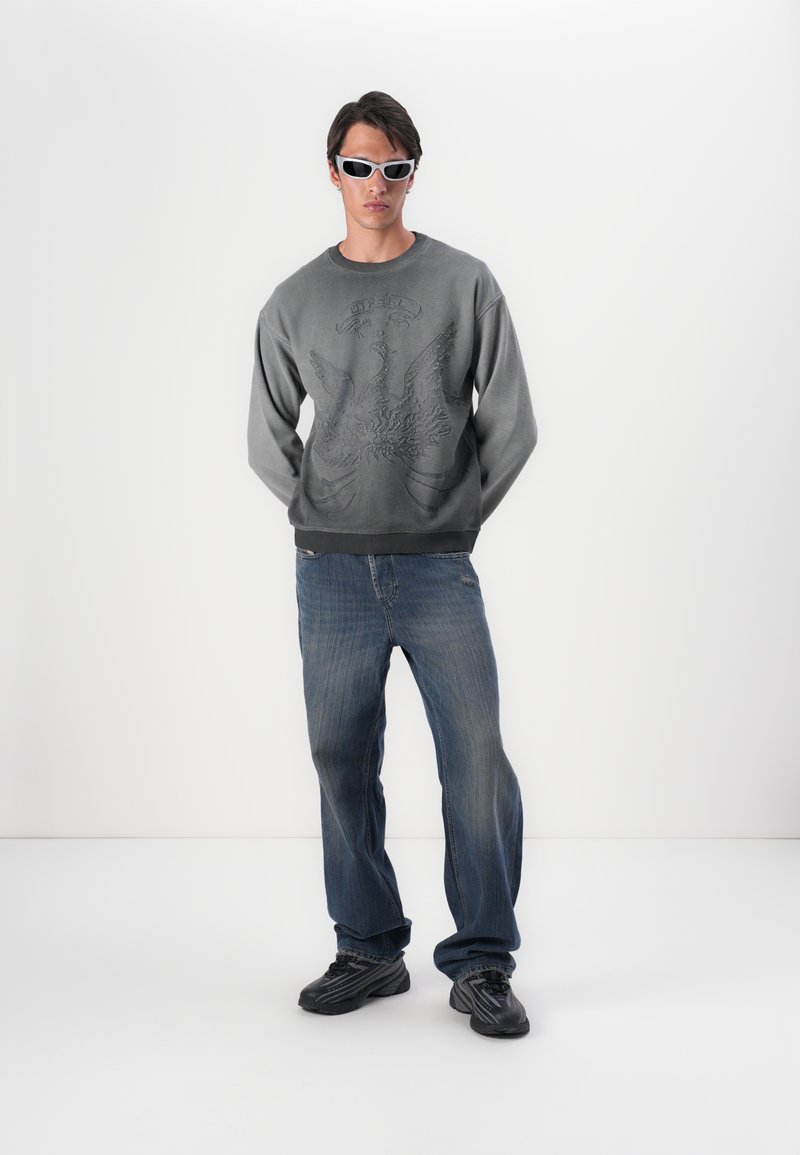 Grå sweatshirt med præget ørnedesign, parret med blå jeans. Modellen bærer hvide solbriller og sorte sneakers. Afslappet sæt.